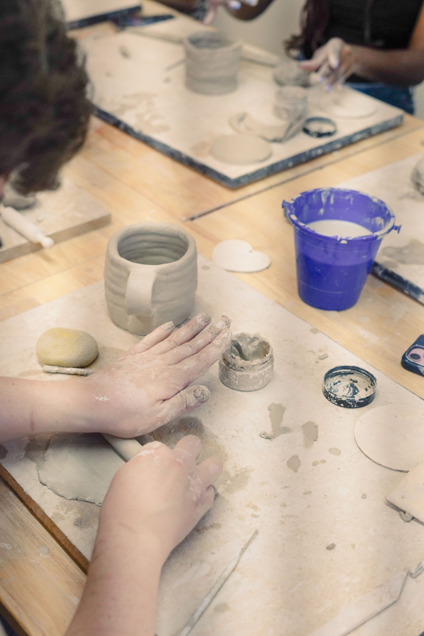 Two hands rolling a coil of clay on a worktable beside a finished mug