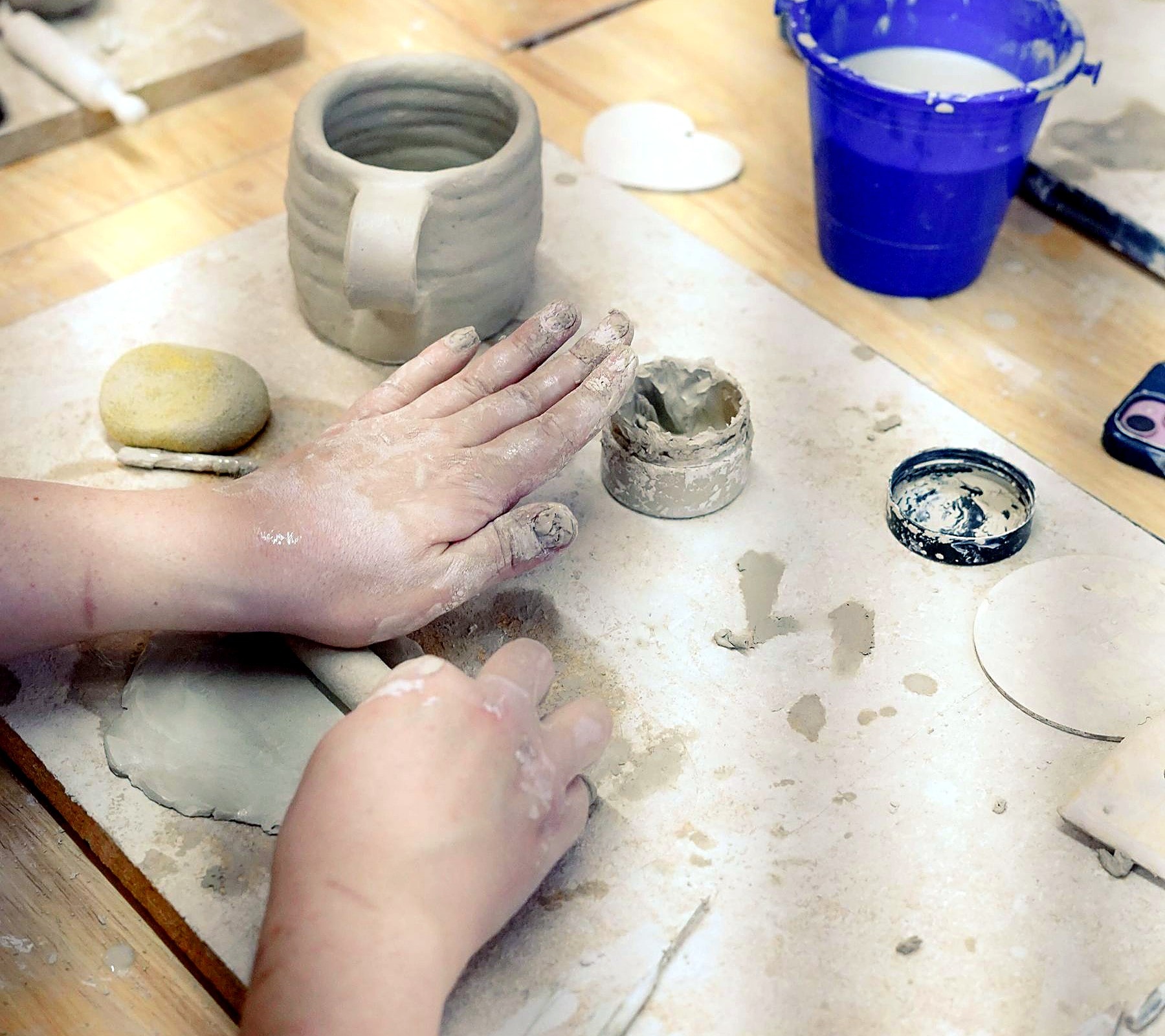 Hands rolling a clay slab beside a handbuilt coil mug on a studio work board